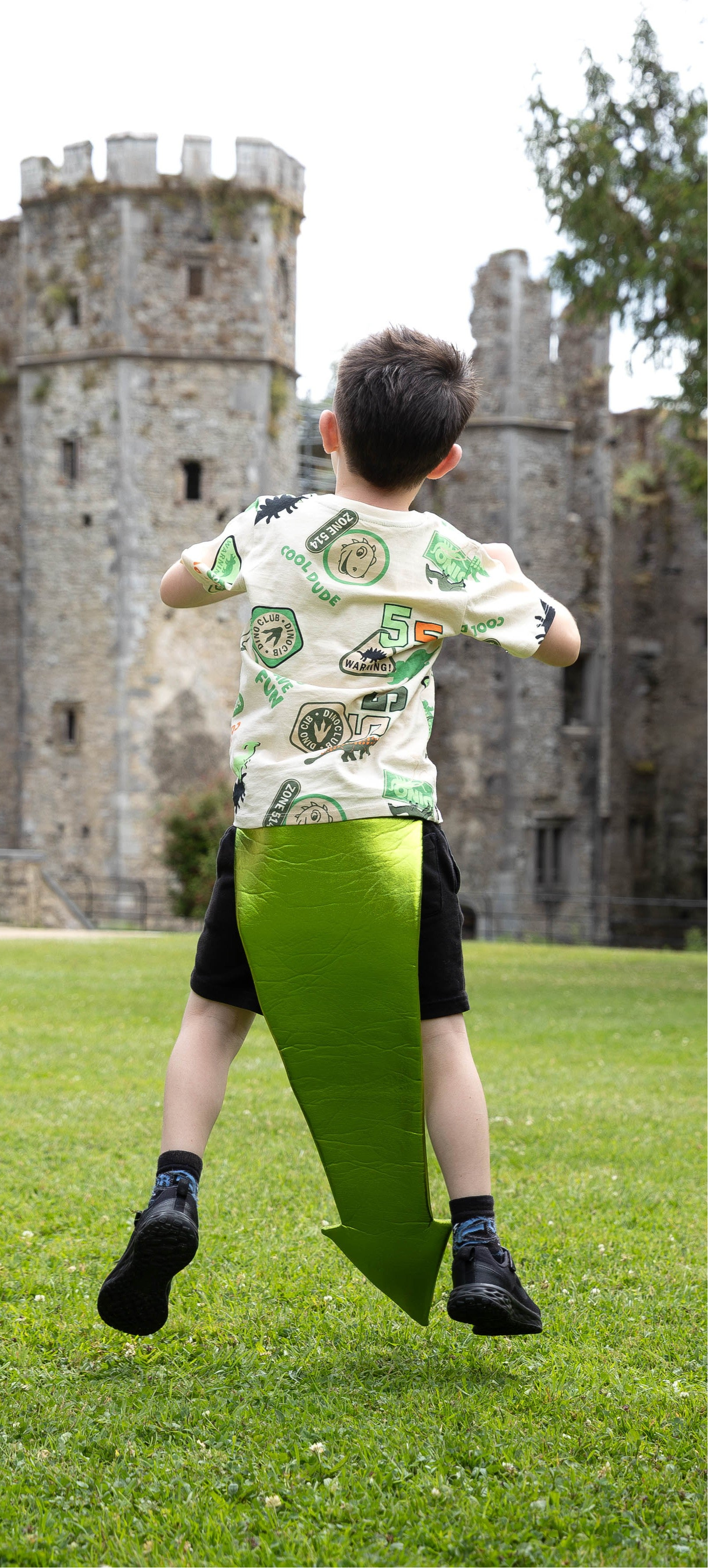 Child wearing a dragon tail, in a patterned shirt and green shorts running on grass with a castle in the background, pretending to be a dragon.