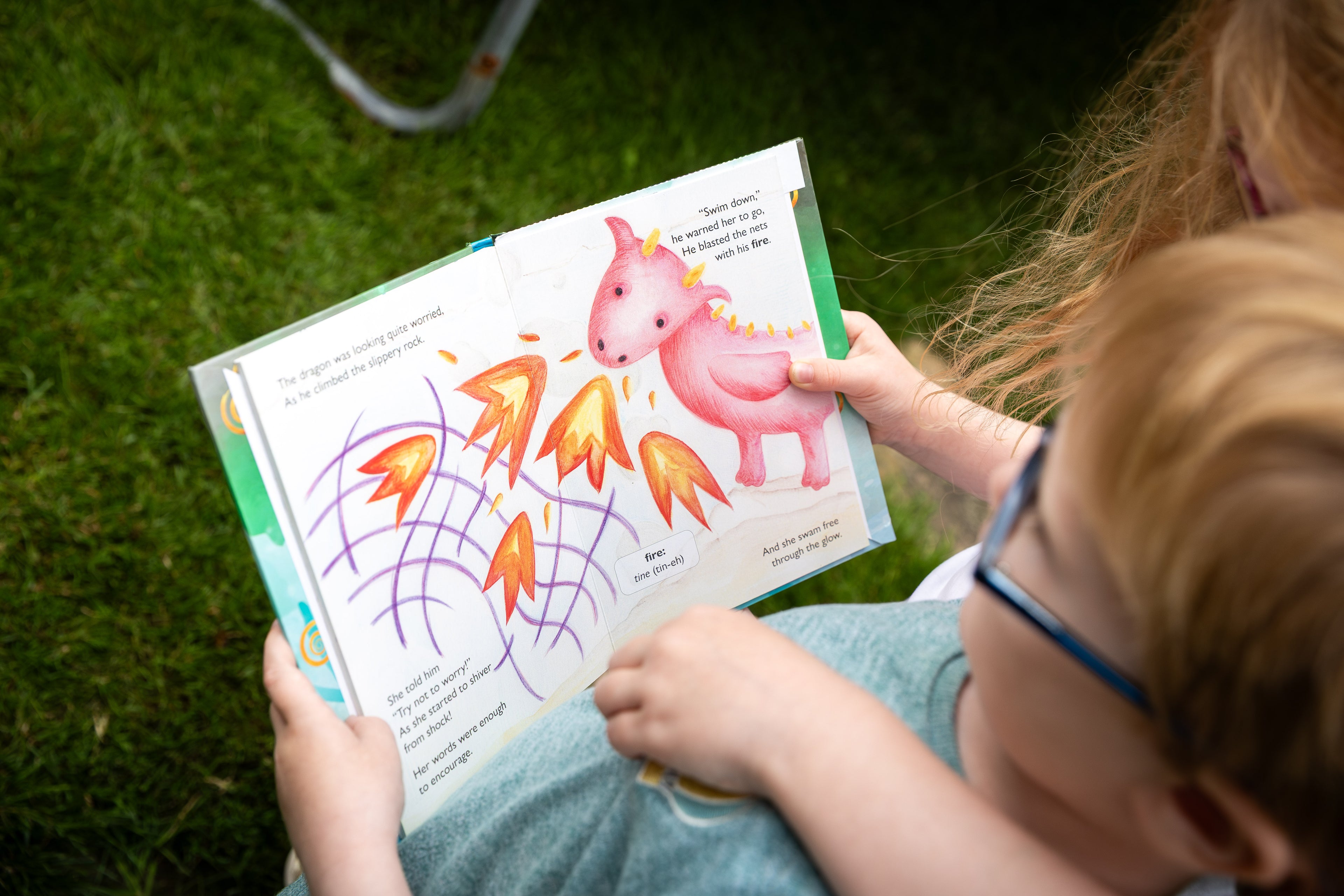 Child reading an Irish Fairy Tails book with a colorful dragon illustration on grass