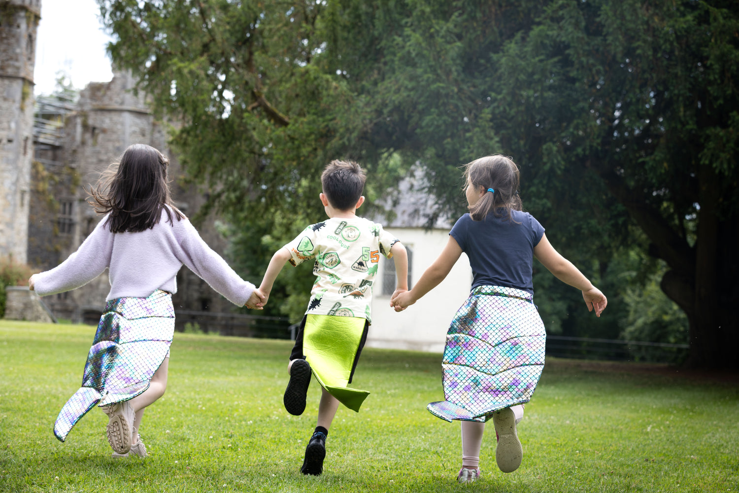 Three children running together wearing dragon tails and mermaid tails in a park with a castle in the background