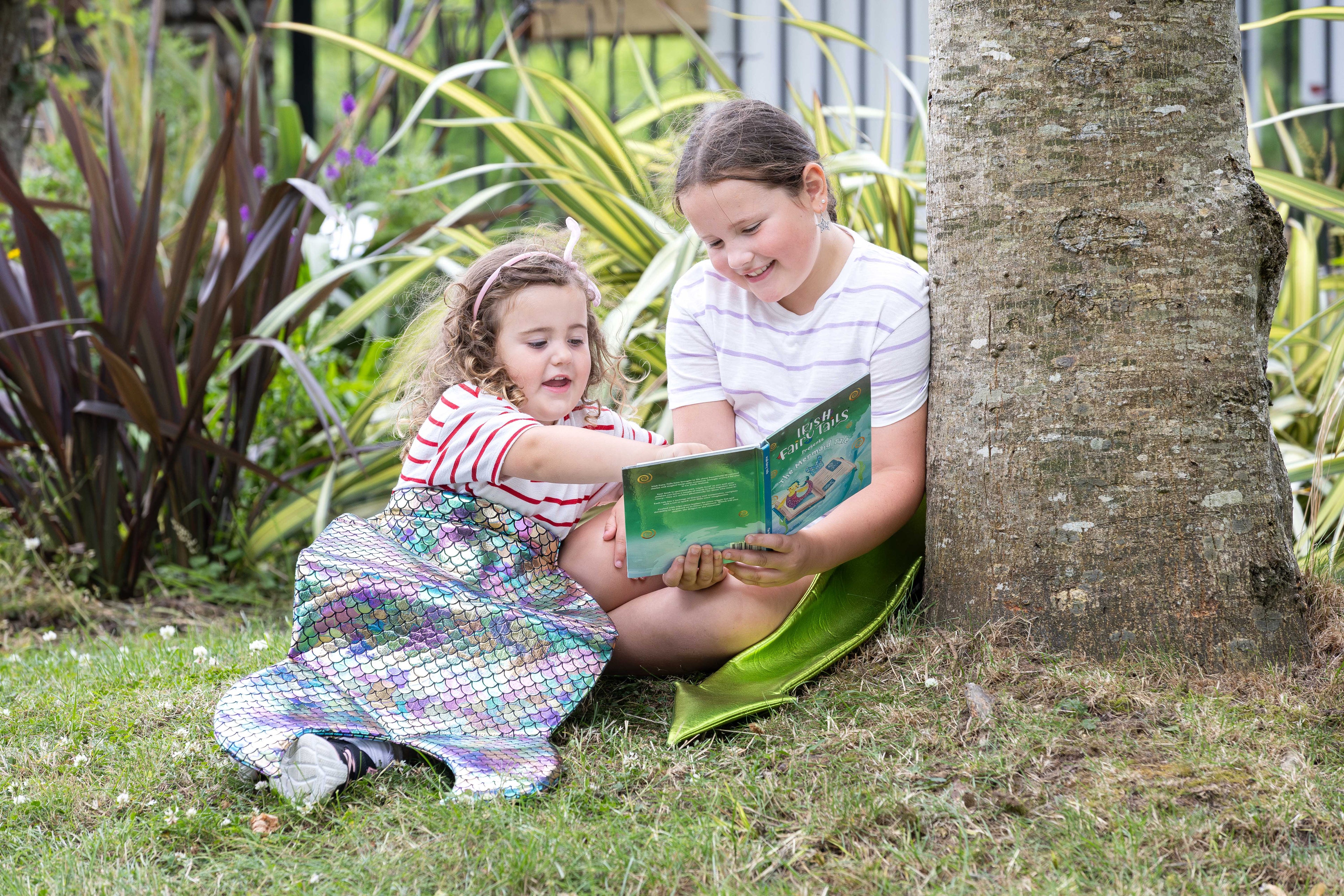 Two young girls reading a book wearing kids costume tails in a garden setting.