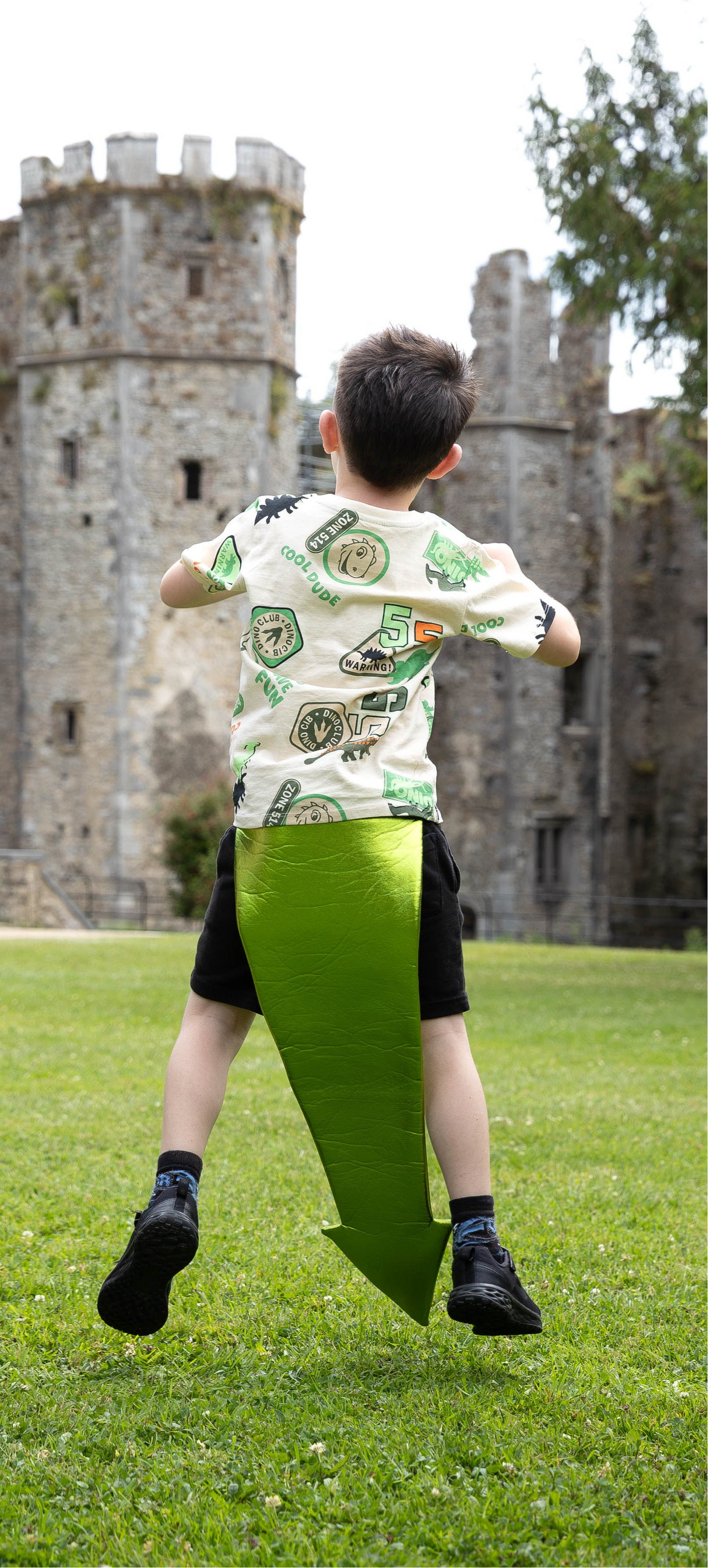 Child wearing a dragon tail, in a patterned shirt and green shorts running on grass with a castle in the background, pretending to be a dragon.