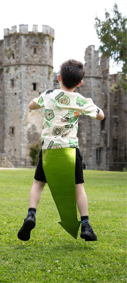 Child wearing a dragon tail, in a patterned shirt and green shorts running on grass with a castle in the background, pretending to be a dragon.
