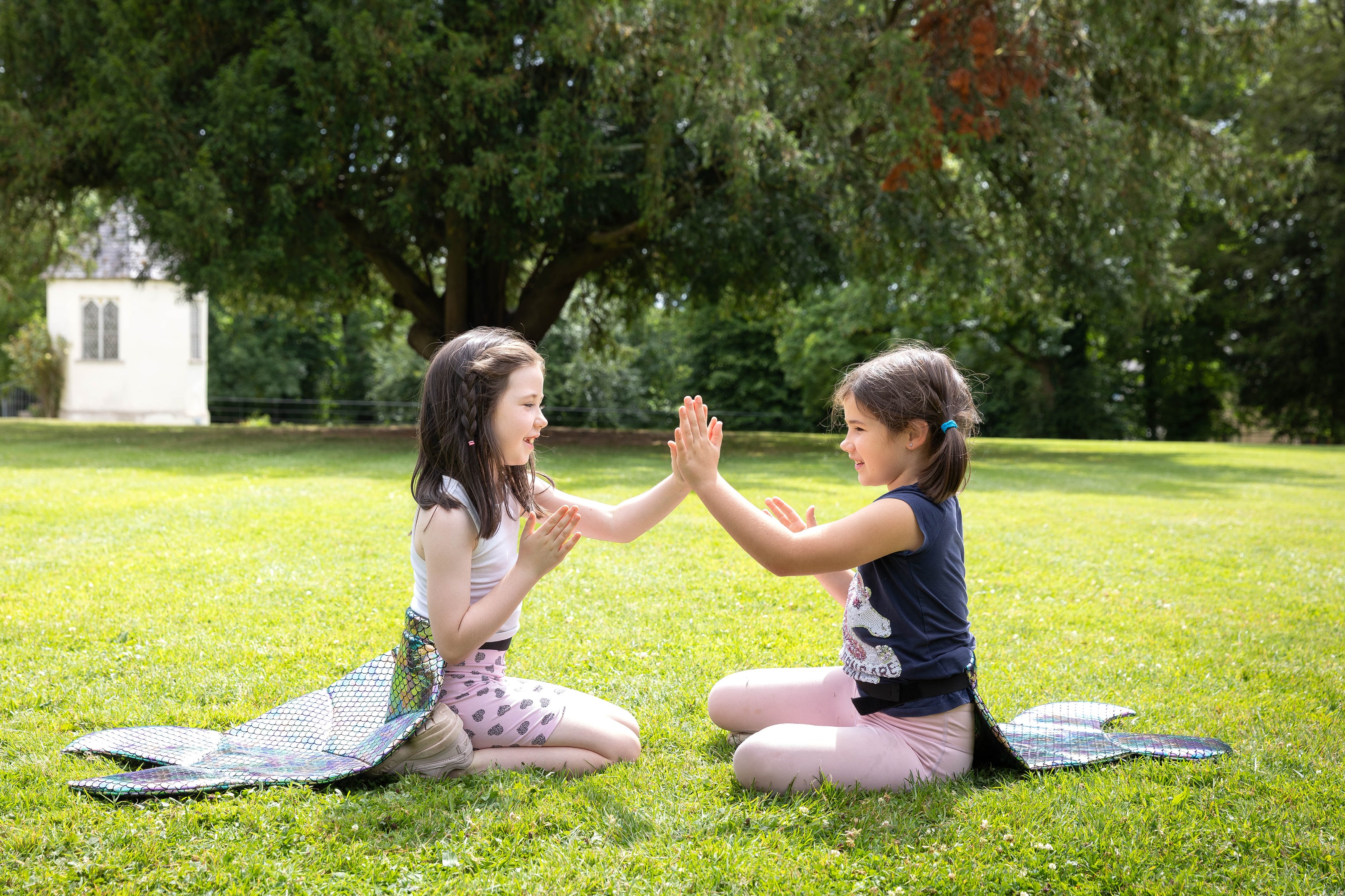 Two young girls wearing mermaid tails in a park, playing.
